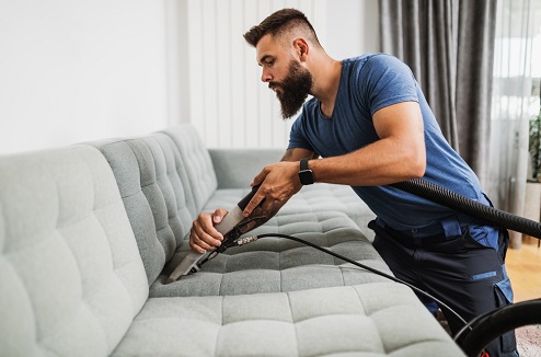 un homme jeune barbu nettoie un canapé gris avec un aspirateur traineau