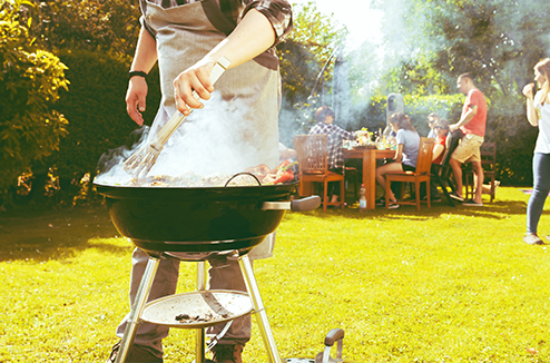 Quel barbecue choisir ? On vous aide Un homme avec un tablier dans un jardin avec des amis, retournant les grillades sur un barbecue à charbon