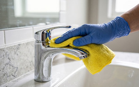 Une main gantée de bleu nettoie un robinet de salle d'eau au chiffon microfibre